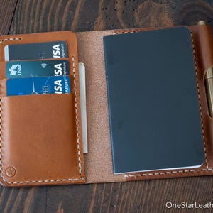 Op de afbeelding: Open bruin leren notitieboek en portemonnee combinatie. Het portemonnee gedeelte bevat meerdere creditcards. Een zwart notitieboek en een pen zijn ook zichtbaar. Het item is gestikt met witte draad.