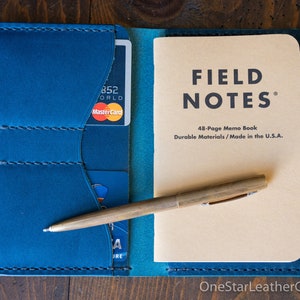 May include: A blue leather wallet with card slots, a Field Notes memo book, and a brass pen. The wallet holds a MasterCard credit card. The memo book reads "FIELD NOTES" and "48-Page Memo Book Durable Materials / Made in the U.S.A."