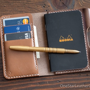 Op de afbeelding: Een bruine leren portemonnee met een gestikte rand, met creditcards, een pen en een klein zwart notitieboekje met het woord "RHODIA" erop gedrukt.