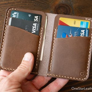 May include: A brown leather card holder wallet featuring two interior pockets with visible stitching along the edges. The wallet contains multiple credit cards, including a black Visa card and a blue Visa card. A small star logo is embossed on the bottom right corner.