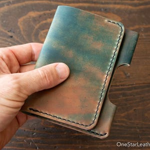 Op de afbeelding: Een opvouwbare lederen portemonnee met een gevlekte blauwgroene en bruine afwerking en zichtbare donkere stiksels langs de randen. De portemonnee heeft een gestructureerd ontwerp met een zijdelingse uitsparing en de tekst OneStarLeatherGoods.com.