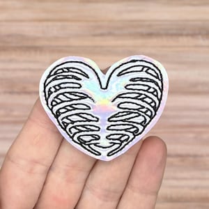 May include: A heart-shaped sticker with a black rib cage design. The sticker has a holographic, iridescent finish that reflects multiple colors. The edges of the sticker are white, and the design is centered.