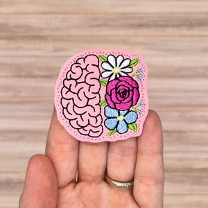 May include: A light pink embroidered patch featuring a stylized human brain on the left side and a cluster of flowers on the right side. The floral arrangement includes a white daisy, a vibrant pink rose, and small light blue blossoms with green leaves.