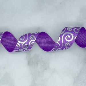 May include: A purple grosgrain ribbon featuring a repeating metallic silver swirl pattern. The ribbon is arranged in a series of horizontal loops, showcasing the textured fabric surface and the reflective quality of the silver printed design.