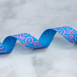 May include: A length of bright blue grosgrain ribbon featuring a repeating pattern of vibrant pink swirls. The ribbon is twisted into a gentle wave shape, showcasing the textured fabric surface and the consistent graphic design printed on the material.