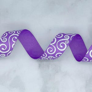 May include: A purple grosgrain ribbon featuring a repeating silver glitter swirl pattern. The ribbon is arranged in a series of horizontal waves, showcasing the textured ribbed fabric and the metallic finish of the decorative spiral design.