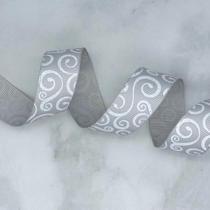 May include: A silver grey grosgrain ribbon featuring a repeating white printed pattern of swirling scrolls and flourishes. The ribbon is folded into a series of soft, undulating curves, showcasing the textured ribbed surface of the fabric.