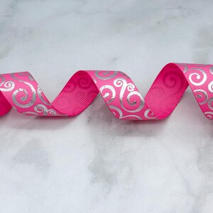 May include: A bright hot pink grosgrain ribbon featuring a repeating metallic silver swirl pattern. The ribbon is arranged in a series of horizontal waves, showcasing the textured ribbed fabric and the shiny, reflective quality of the silver print.