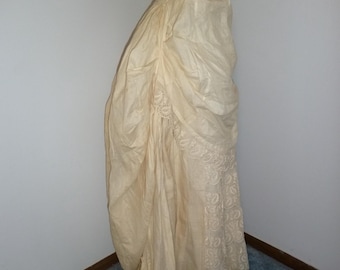May include: A cream-coloured vintage skirt with detailed lace edging. The skirt is draped and gathered, creating a flowing silhouette. It is displayed on a black mannequin, against a neutral wall and blue carpet.