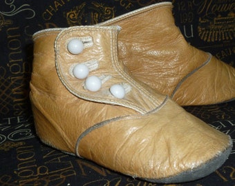 May include: A pair of tan leather baby shoes with white button closures. The shoes have a rounded toe and a low heel.