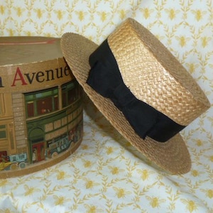 May include: A straw boater hat with a black grosgrain ribbon band rests beside a vintage hat box. The hat box is cylindrical and features an illustration of a building with the words "Fifth Avenue" on the top.
