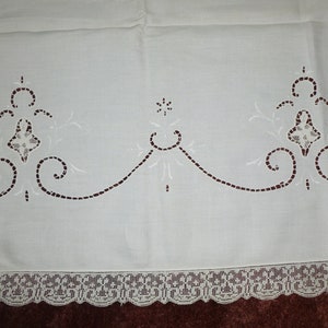 K&ouml;nnte beinhalten: Wei&szlig;e bestickte Tischdecke mit zarter Spitzenborte. Die Tischdecke zeigt ein sich wiederholendes Muster aus floralen Motiven und Schn&ouml;rkeln.