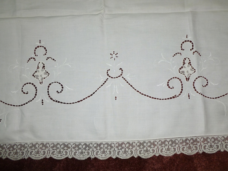 K&ouml;nnte beinhalten: Wei&szlig;e bestickte Tischdecke mit zarter Spitzenborte. Die Tischdecke zeigt ein sich wiederholendes Muster aus floralen Motiven und Schn&ouml;rkeln.
