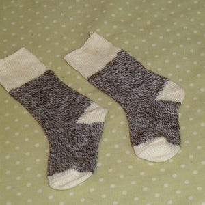 May include: A pair of knit socks featuring a heathered brown and white speckled pattern on the main body, with solid cream-colored cuffs, heels, and toes. The socks are constructed with a ribbed texture at the top and a smooth knit finish.