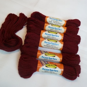May include: A collection of burgundy Persian type yarn skeins. Five skeins are neatly stacked, each with a label. A separate bundle of yarn sits to the left. The labels read "Persian Type Yarn" and "100% wool for needlepoint and crewel."