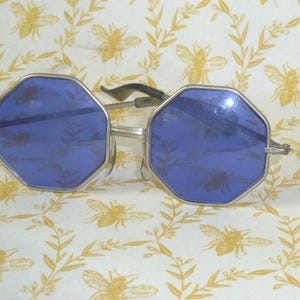 May include: Silver-framed sunglasses with octagonal blue lenses. The lenses have a subtle pattern. The arms are black and silver. The background is a cream-colored fabric with a repeating pattern of bees and leaves.