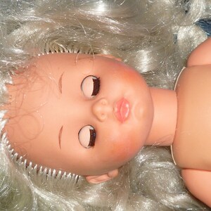 Puede incluir: Una mu&ntilde;eca con el pelo largo y rubio y los ojos cerrados. La mu&ntilde;eca tiene la boca rosa y lleva un vestido blanco.