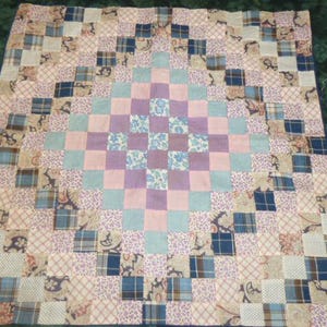 May include: A patchwork quilt with a central diamond pattern. The quilt features squares of various fabrics in shades of pink, blue, purple, and cream. The quilt's dimensions are not visible.