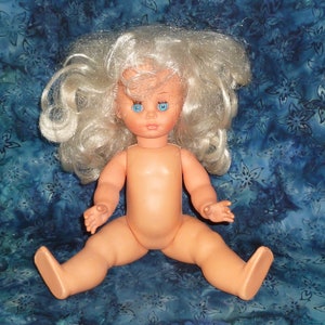 Puede incluir: Una mu&ntilde;eca de pl&aacute;stico desnuda con el pelo largo, blanco y rizado y ojos azules. La mu&ntilde;eca est&aacute; sentada sobre una tela de estampado azul y blanco.