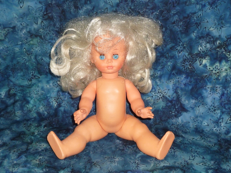 Puede incluir: Una mu&ntilde;eca de pl&aacute;stico desnuda con el pelo largo, blanco y rizado y ojos azules. La mu&ntilde;eca est&aacute; sentada sobre una tela de estampado azul y blanco.