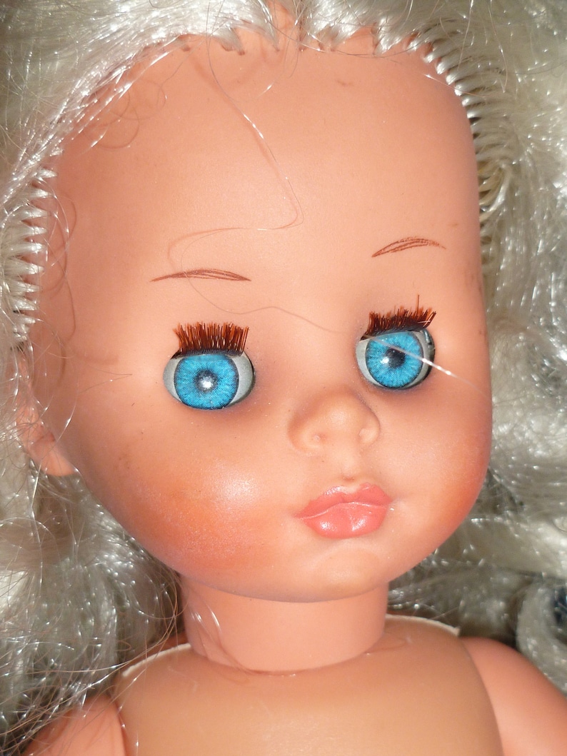 Puede incluir: Una mu&ntilde;eca de porcelana con el pelo largo y blanco y ojos azules. La mu&ntilde;eca tiene una sonrisa pintada y mejillas sonrosadas.