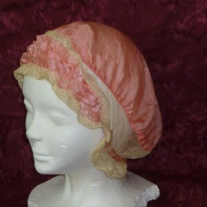 Puede incluir: Una gorro de dormir de satén rosa con un borde de encaje. El gorro está fruncido en la parte superior y tiene un borde rizado.