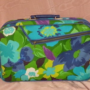 May include: Vintage floral suitcase with a vibrant pattern of blue, green, purple, and yellow flowers. The suitcase has a blue handle, trim, and zipper. The bag is rectangular and has a zippered front pocket.