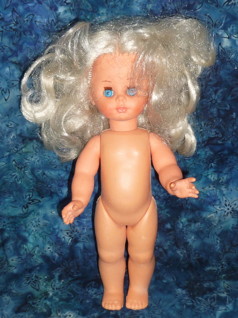 Puede incluir: Una mu&ntilde;eca vintage con el pelo largo, blanco y rizado. La mu&ntilde;eca tiene ojos azules y no lleva ropa. La mu&ntilde;eca est&aacute; de pie sobre una superficie con un patr&oacute;n azul y blanco.