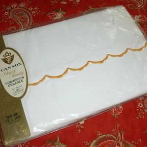 Può includere: Un lenzuolo bianco per letto singolo in una confezione di plastica con un'etichetta dorata. L'etichetta dice "Cannon Royal Family Combspun Percale" e "Twin Bed 72 x 108". Il lenzuolo ha un bordo smerlato dorato.
