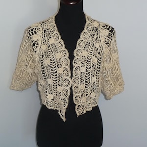 Może przedstawiać: Kremowy, szydełkowy bolerko z otwartym przodem i krótkimi rękawami. Delikatny wzór koronki przedstawia wzory kwiatowe i geometryczne. Bolerko jest prezentowane na czarnym manekinie, prezentując jego skomplikowane detale i fakturę.