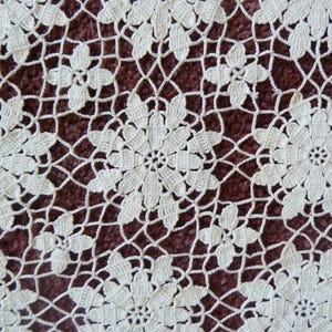 May include: Close-up of a cream-colored, crocheted doily with a floral pattern. The design features large, central flowers surrounded by smaller blossoms and a delicate, openwork background. The doily is set against a dark, reddish-brown surface.