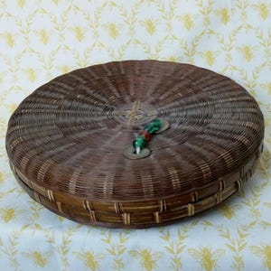 以下が含まれることがあります： 茶色の柳で作られた、蓋付きの丸い編みかご。 かごには留め金があり、緑と赤のビーズで飾られています。 黄色の蜂の模様が入った白い表面に置かれています。 直径約30cm。