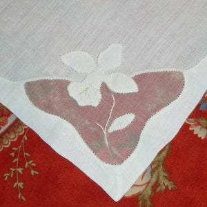 May include: A white linen napkin with a delicate embroidered flower design. The embroidery is in white thread and features a flower with five petals and a stem with leaves. The napkin is shown against a red background with a floral pattern.