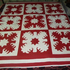 May include: A red and white quilt with a repeating floral pattern. The quilt features large squares with a central star design surrounded by stylized floral shapes. The quilt is on a bed, showcasing its size and design.
