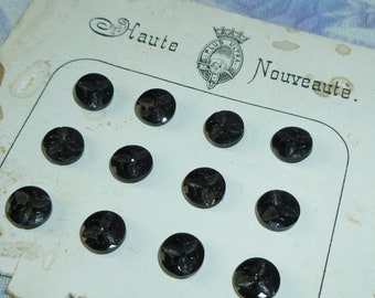 May include: A card with the text "Haute Nouveauté" and a crest with the text "Plus Ultra" in the centre. The card has 12 black glass buttons with a floral design arranged in rows.