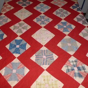 May include: A red quilt with a diamond pattern. Each diamond is filled with a patchwork design of blue, pink, and white fabric. The quilt is made of various fabrics, including plaid, striped, and floral patterns.