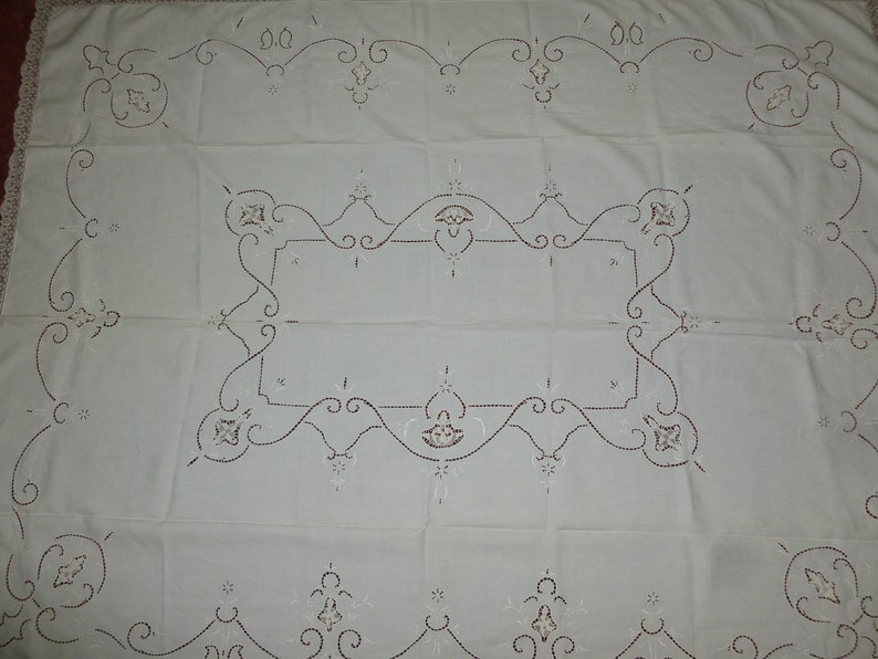 K&ouml;nnte beinhalten: Wei&szlig;e bestickte Tischdecke mit einem zarten Blumenmuster. Die Tischdecke ist quadratisch und hat ein zentrales Design mit einem Rand aus komplizierten Schn&ouml;rkeln.