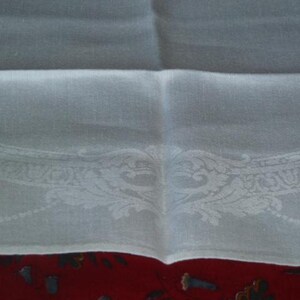 Op de afbeelding: Wit linnen tafelkleed met een delicate geborduurde rand met een bloemenmotief. Het tafelkleed is dubbelgevouwen, waardoor de ingewikkelde details van de borduursels zichtbaar worden.