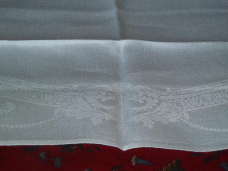 Op de afbeelding: Wit linnen tafelkleed met een delicate geborduurde rand met een bloemenmotief. Het tafelkleed is dubbelgevouwen, waardoor de ingewikkelde details van de borduursels zichtbaar worden.