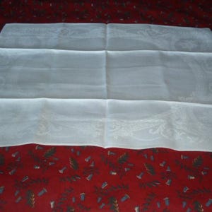 Op de afbeelding: Een wit linnen tafelkleed met een delicaat geborduurd ontwerp. Het tafelkleed is dubbelgevouwen en ligt op een rode achtergrond.