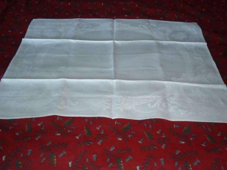 Op de afbeelding: Een wit linnen tafelkleed met een delicaat geborduurd ontwerp. Het tafelkleed is dubbelgevouwen en ligt op een rode achtergrond.