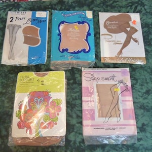 May include: Five packages of vintage hosiery are displayed on a green surface. The packages are in various colors, including blue, white, pink, and yellow. The packages feature illustrations of legs and floral designs, with text such as "Seamless Mesh" and "Total Support".
