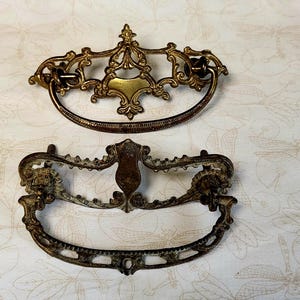 May include: Two antique brass drawer pulls with ornate designs. The top pull has a more intricate design with a central oval shape. The bottom pull has a more simple design with a central oval shape.