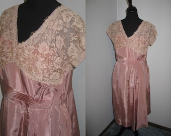 May include: A dusty rose knee-length dress featuring a V-neckline and short sleeves. The bodice is adorned with an overlay of cream-colored floral lace. The waist is accented with a wide, pleated satin sash that ties at the side, creating a draped effect.