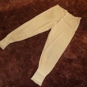 May include: A pair of cream-coloured long johns featuring a button-front closure at the waist. The garment is constructed with ribbed cuffs at the ankles and a fitted waistband, showcasing a simple, functional design with a light, neutral tone.
