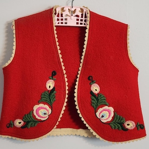May include: A red wool vest with floral embroidery. The vest has a cream-coloured scalloped trim and is decorated with embroidered flowers in shades of pink, green, and white. The vest is hanging on a pink hanger.