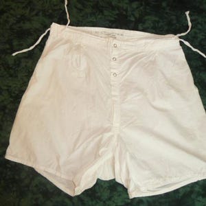 May include: White cotton shorts with a button-up front closure and drawstring ties at the waist. The shorts have a relaxed fit and are made of a lightweight fabric. The garment is likely vintage or antique.