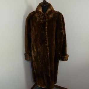 May include: A long, brown faux fur coat with a wide collar and button closures. The coat is displayed on a mannequin, showcasing its full length and textured appearance. The sleeves have turned-up cuffs.