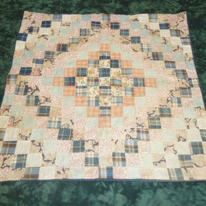 May include: A patchwork quilt with a central diamond pattern. The quilt features squares of various fabrics in shades of blue, peach, and cream. The design includes floral and plaid patterns, creating a cozy, handcrafted aesthetic. The quilt is a square shape.