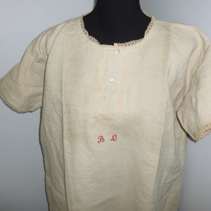 May include: A cream-colored linen shirt with short sleeves and a lace trim around the neckline and sleeves. The shirt has a button-up front and the initials "B L" embroidered on the front in red thread.